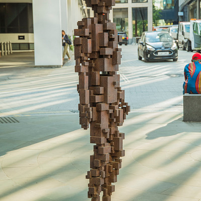 The image features a large, intricately constructed wooden statue of a man standing on the sidewalk. This unique sculpture is made up of several blocks of wood and appears to be an artistic representation of a person. The scene takes place in a busy city area with multiple vehicles present.