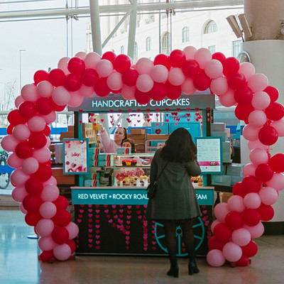 The image features a woman standing in front of a cake shop, which has a unique and eye-catching display. A large heart made out of balloons is prominently displayed outside the store, drawing attention to the shop's offerings. There are several other people around the area, some closer to the woman and others further away.