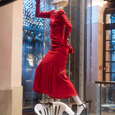 The image features a mannequin dressed in a red dress, standing on top of a white chair. The mannequin appears to be posing for the camera, with its legs crossed and hands placed on the chair's back. The scene takes place inside a store or showroom, as evidenced by the presence of multiple chairs scattered throughout the space.