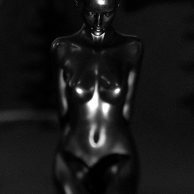 The image is a black and white photo of a naked woman statue. She appears to be sitting on a chair, with her legs crossed. The statue has an artistic and elegant appearance, capturing the essence of a female form.
