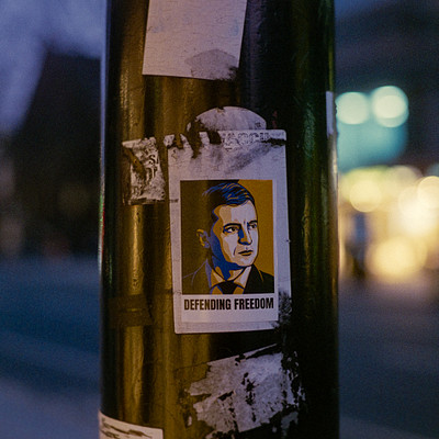 The image features a black pole with various stickers on it, including one that says "Defending Freedom." There are also two other stickers on the pole. In addition to these stickers, there is a sticker of a man's face placed above them. The scene takes place at night, as evidenced by the darkness surrounding the pole and the streetlights in the background.