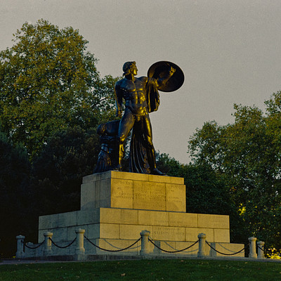 The image features a statue of a man holding a shield, standing on top of a pedestal. The statue is located in the middle of a park or field, surrounded by trees and bushes. There are several benches placed around the area, providing seating for visitors to enjoy the view of the statue.