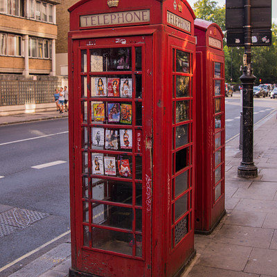 The image features two red telephone booths, also known as red phone boxes or red post offices, located on a sidewalk. They are placed next to each other and appear to be vending machines for DVDs. There is a variety of DVD covers displayed in the windows of these booths, showcasing different movies available for purchase.