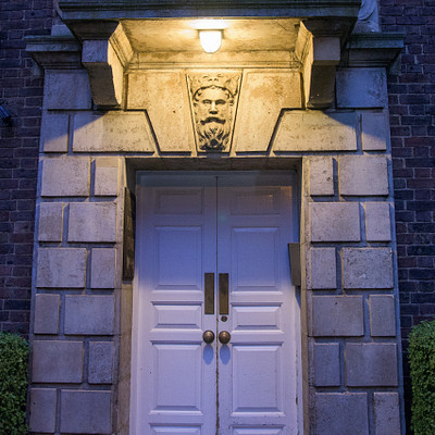 The image features a large, old-fashioned building with a white door and black trim. Above the doorway, there is a light fixture that illuminates the entrance, creating an inviting atmosphere. The building appears to be made of brick, giving it a classic and timeless appearance.