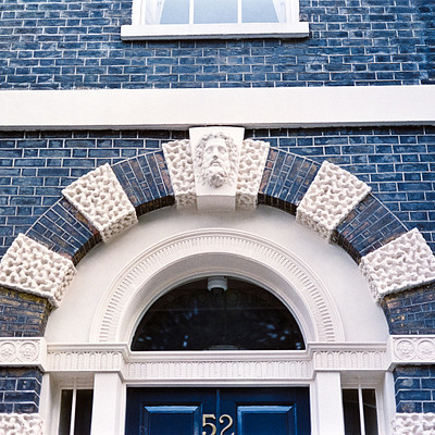 The image features a large, ornate building with a blue brick facade. A prominent archway is adorned with intricate carvings and decorative elements, including a stone face above the door. The entrance to the building has a black door with gold numbers on it, adding an elegant touch to the overall design.
