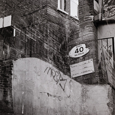 The image is a black and white photo of an old brick building with graffiti on the side. There are two signs attached to the wall, one of which reads "SPC 40." Above these signs, there is a window that provides natural lighting to the scene. Additionally, there is a clock mounted on the wall near the top right corner of the image.