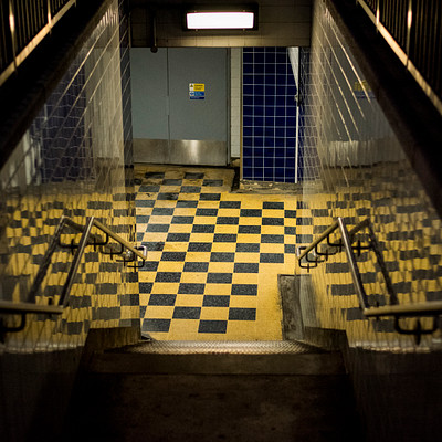 The image is a black and white photo of an outdoor stairway with yellow and blue checkered tiles. There are two sets of stairs, one on the left side and another on the right side of the image. A metal railing can be seen in front of the stairs, providing safety for those using them.