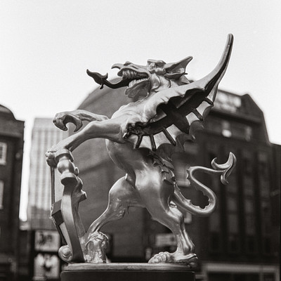 The image is a black and white photo of a statue of a dragon, which appears to be made of metal. It has its mouth open as if it's roaring or making a sound. The statue is located in the middle of a city street, surrounded by buildings on both sides.