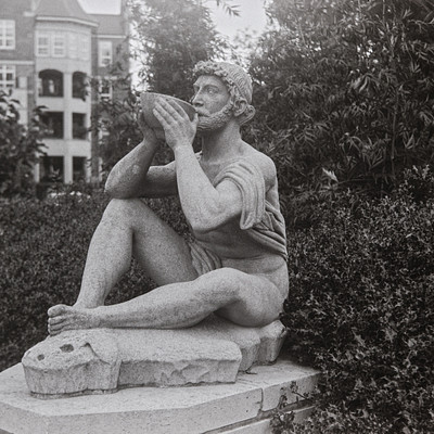 The image is a black and white photograph of a statue, possibly depicting a man or a woman. The statue appears to be sitting on a stone pedestal in an outdoor setting, surrounded by greenery. It seems to be a public art piece, drawing attention from passersby.