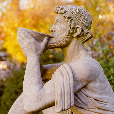 The image features a statue of a man holding a cup in his hand. He appears to be drinking from the cup, possibly enjoying a beverage or observing something around him. The statue is positioned outdoors and seems to be part of an artistic display or garden setting.