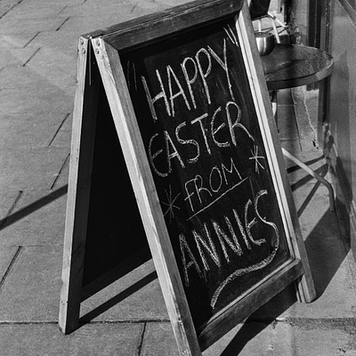 The image is a black and white photo of an outdoor scene featuring a chalkboard sign that reads "Happy Easter from Annies." The sign is placed on the sidewalk, possibly in front of a store or restaurant. There are two chairs visible in the background, one located near the left edge of the image and another towards the right side. Additionally, there is a dining table situated further back in the scene.