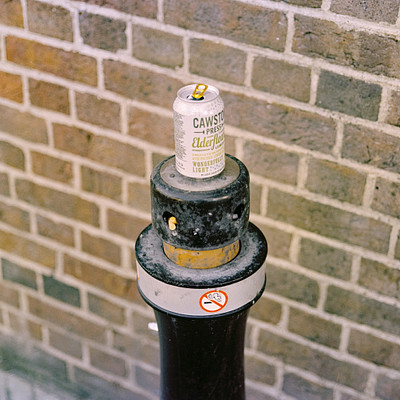 The image features a parking meter with a can of beer on top. The can is placed in the middle of the meter, and it appears to be an empty can. The scene takes place outdoors, possibly near a brick wall or building.
