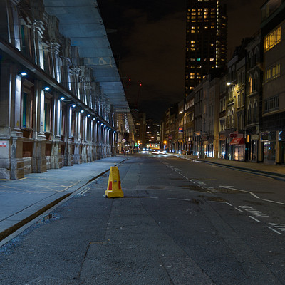 The image is a black and white photo of an empty city street at night. There are several people walking along the sidewalk, with some closer to the foreground and others further away. A few cars can be seen parked or driving on the street, adding to the urban atmosphere.