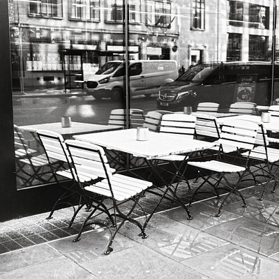 The image is a black and white photo of an outdoor cafe with several tables and chairs arranged outside. There are at least 12 chairs visible in the scene, some placed near the dining tables while others are scattered around the area. A few benches can also be seen among the seating options.