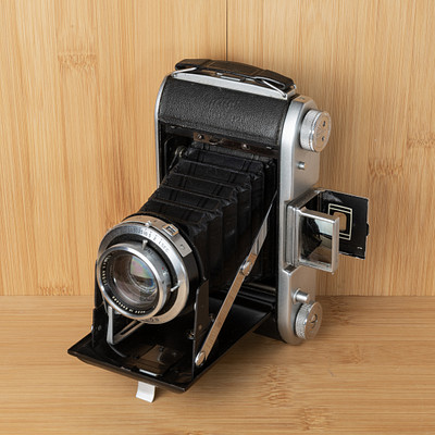 The image features an old-fashioned camera sitting on a wooden table. It is placed in the center of the scene, with its lens facing upwards. The camera appears to be black and silver, giving it a vintage appearance.