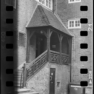 The image is a black and white photo of an old brick building with a steeple. The building has a large porch, which features a staircase leading up to the entrance. There are two people standing in front of the building, one closer to the left side and another nearer to the right side.