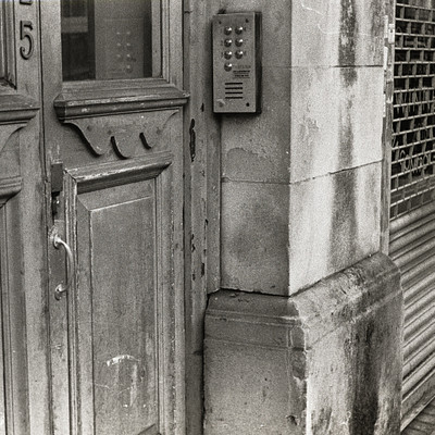 The image is a black and white photo of an old building with a doorway. A telephone booth is located next to the door, which has a dial pad on it. There are two doors in the scene; one is open while the other is closed.