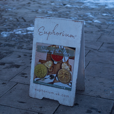 The image is a black and white photo of a sign on the sidewalk. The sign has a picture of food, including apples and oranges, and says "Euphorium" in large letters. It appears to be advertising something related to food or dining.