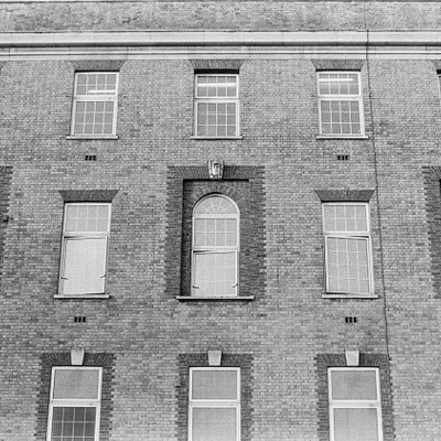 The image is a black and white photo of an old brick building with many windows. There are at least 14 visible windows, each varying in size and position on the building's facade. Some windows have shutters or blinds covering them, adding to the overall architectural charm of the structure.