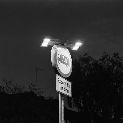 The image is a black and white photo of a street sign with a bus symbol on it. The sign is located near the side of a road, possibly in an urban setting. There are trees visible in the background, adding to the atmosphere of the scene.