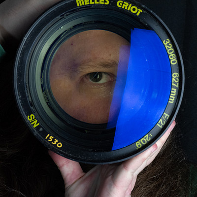 The image shows a close-up of a person's face, with the camera lens covering most of the frame. The person is looking through the viewfinder of a camera, capturing their own reflection in the lens.