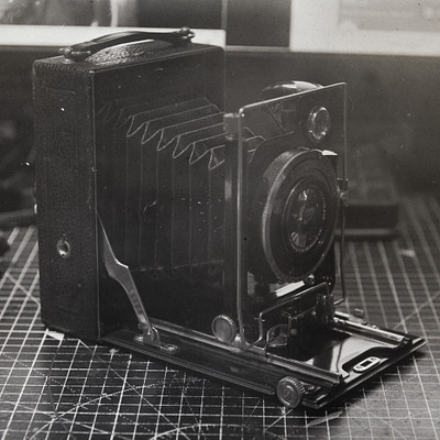 The image is a black and white photograph of an old-fashioned camera. It appears to be a vintage model, possibly from the 1950s or earlier. The camera has a large lens on top and is placed on a table.