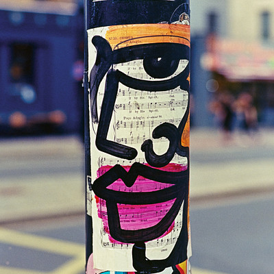 The image features a pole with various stickers on it, including one that has an eye and mouth drawn on it. There are also other stickers covering the pole, giving it a colorful appearance. In the background, there is a street scene with several people walking around.