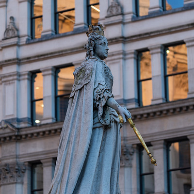 The image features a statue of a woman, possibly Queen Victoria, sitting on top of a pedestal. She is holding a scepter in her hand and appears to be wearing a crown. The statue is located outside of a building with many windows, giving the impression that it might be situated in a city square or public area.
