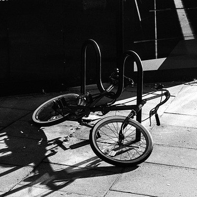The image is a black and white photo of an old bicycle parked next to a bike rack. The bicycle is leaning against the rack, which has two other bikes on it. There are also three benches in the scene, one located near the left side of the frame, another in the middle, and the third one towards the right side.