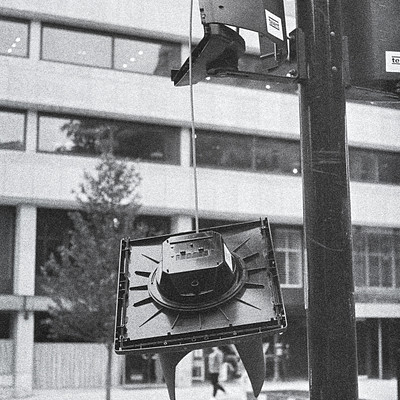 The image is a black and white photo of an old television hanging from a traffic light. It appears to be a creative or artistic display, as the TV is not typically hung in such a manner. In addition to the unusual sight of the television, there are several people walking around the area.