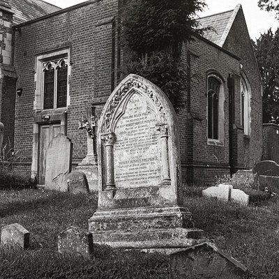 The image is a black and white photo of an old cemetery with several headstones. A prominent headstone stands out, featuring a cross on top and the words "In Loving Memory" written below it. This particular headstone is located in front of a brick building, possibly a church or a historical site.