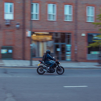 The image captures a man riding a motorcycle down the street, passing by a building. He is wearing a helmet for safety while enjoying his ride. There are several other people in the scene, some of them standing or walking around, and others sitting on benches.