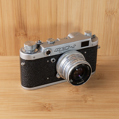 The image features an old-fashioned camera sitting on a wooden table. The camera is silver and black in color, with its lens facing the viewer. It appears to be an antique model, possibly a vintage camera from the 1950s or earlier.