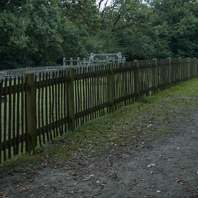 The image features a dirt road that runs through a fenced-in area. Along the path, there are several benches placed at regular intervals for people to sit and enjoy the surroundings. Some of these benches are located closer to the fence while others are further away from it.