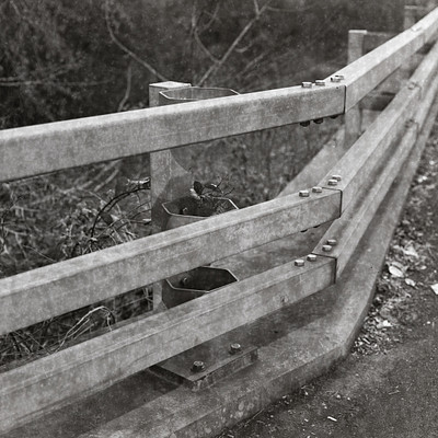 The image is a black and white photo of a metal guard rail on the side of a road. The guard rail appears to be broken, with one section hanging downwards. There are several bolts visible in the rail, which seem to have been loosened or removed.