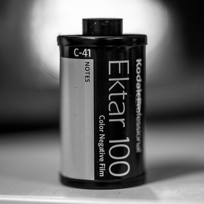 The image features a close-up of a Kodak Ektar 100 film roll sitting on top of a table. The film is black and silver, with the word "Ektar" written in white letters on it. The focus of the photo is on the film itself, highlighting its color negative properties.