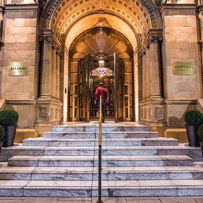 The image features a large, ornate building with an impressive staircase leading up to its entrance. A person is standing at the top of the stairs, possibly waiting for someone or preparing to enter the building. There are several potted plants placed around the area, adding a touch of greenery and enhancing the overall ambiance.
