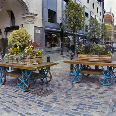 The image depicts a bustling city street with several people walking around. There are two wooden carts, one blue and the other red, parked on the sidewalk. These carts are filled with various potted plants, creating an attractive display for passersby to admire.