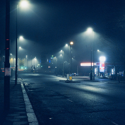 The image depicts a dark city street at night, with the foggy atmosphere creating an eerie ambiance. There are several cars parked along the side of the road, and some traffic lights can be seen in various locations throughout the scene. A few people are walking on the sidewalk, adding to the sense of activity despite the late hour.