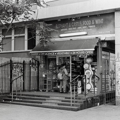 The image is a black and white photo of an outdoor market with a man standing in front of it. There are several people around the area, some walking by and others browsing through the market. A person can be seen carrying a handbag as they walk past the storefront.