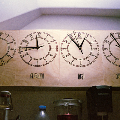 The image features a kitchen with a wooden clock hanging on the wall. This clock is unique as it has four faces, each displaying different times in various languages. There are three bottles placed around the kitchen area, one near the left side of the clock and two others closer to the right side.
