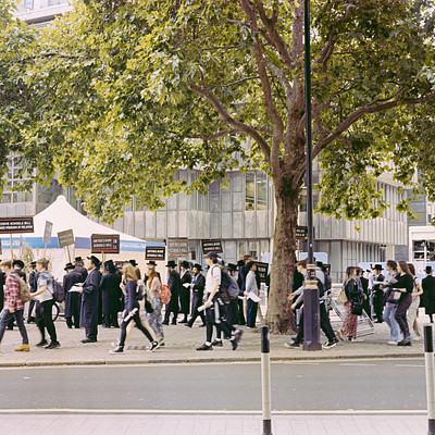 The image is a black and white photo of a large group of people walking down the street. They are all wearing backpacks, with some carrying handbags as well. There are at least 14 people visible in the scene, spread out along the sidewalk.