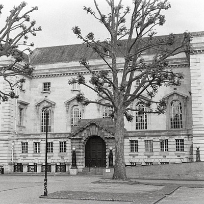 The image is a black and white photo of an old building with a large doorway. There are several trees surrounding the building, providing shade to the area. In front of the building, there are two benches where people can sit and enjoy the surroundings.