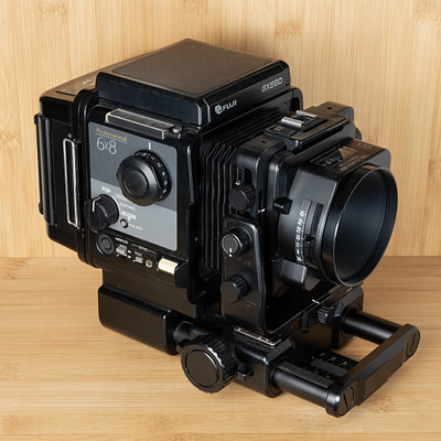 The image features a vintage camera sitting on top of a wooden table. The camera is black and appears to be an old-fashioned model, possibly a Polaroid or a similar brand. It has various buttons and knobs on it, indicating its functionality as a photography device.