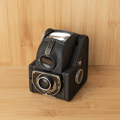 The image features a vintage camera sitting on top of a wooden table. The camera is black and appears to be an old-fashioned model, possibly made by Kodak. It has a leather case that adds to its antique charm.