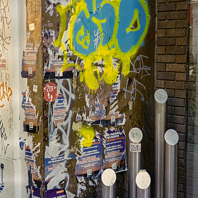 The image features a brick wall with graffiti on it, including a yellow smiley face. There are two parking meters attached to the wall, one closer to the left side and another further right. Above these parking meters, there is a collection of flyers posted on the wall, adding more visual interest to the scene.