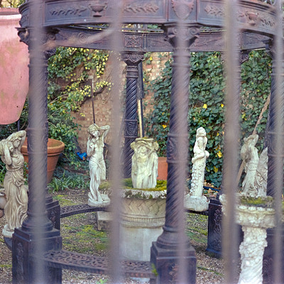 The image features a garden with several statues and vases. There are five statues in total, each depicting different figures. Some of the statues are placed on pedestals or raised platforms, while others are positioned closer to the ground.
