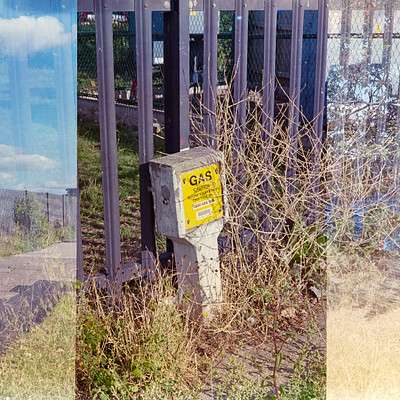 The image is a black and white photo of an outdoor scene with a metal fence in the foreground. There are two signs on the fence, one being a yellow sign that says "gas" and another smaller sign below it. A parking meter can be seen next to the fence, adding to the urban atmosphere of the scene.