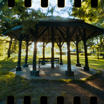 The image features a large wooden pavilion with a shingled roof, situated in the middle of a park. The pavilion has several benches surrounding it, providing ample seating for visitors to enjoy the outdoor space. There are at least five benches visible in the scene, some closer to the pavilion and others further away.