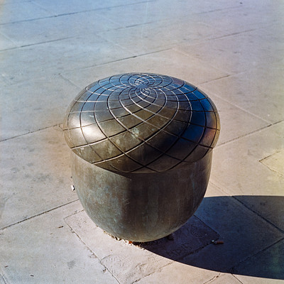 The image features a large, circular metal sculpture sitting on the ground. It appears to be made of metal and has an interesting design with a patterned surface. The sculpture is placed in a concrete area, possibly a sidewalk or a patio.